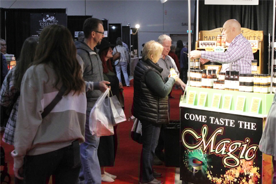 A lively market scene shows a vendor offering samples at a booth labeled "Come Taste the Magic." Customers engage with the vendor while exploring various products on display, surrounded by other stalls in a bustling event space with a vibrant red carpet.