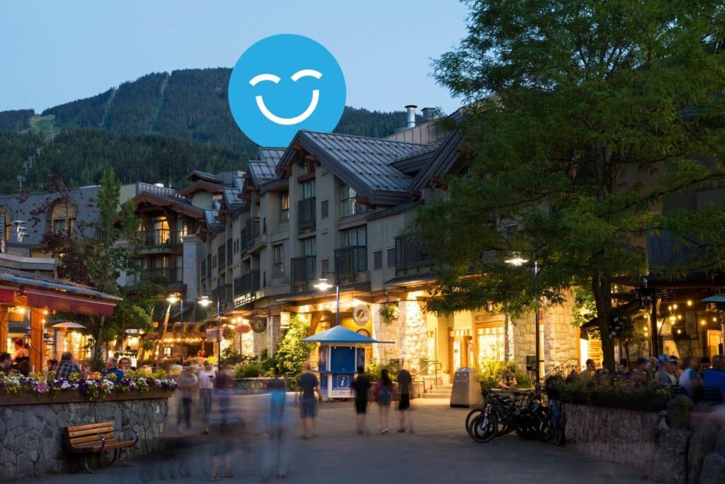 A bustling evening scene in a village with people walking, illuminated shops, and a backdrop of mountains. A blue circle with a smiling face hovers above.