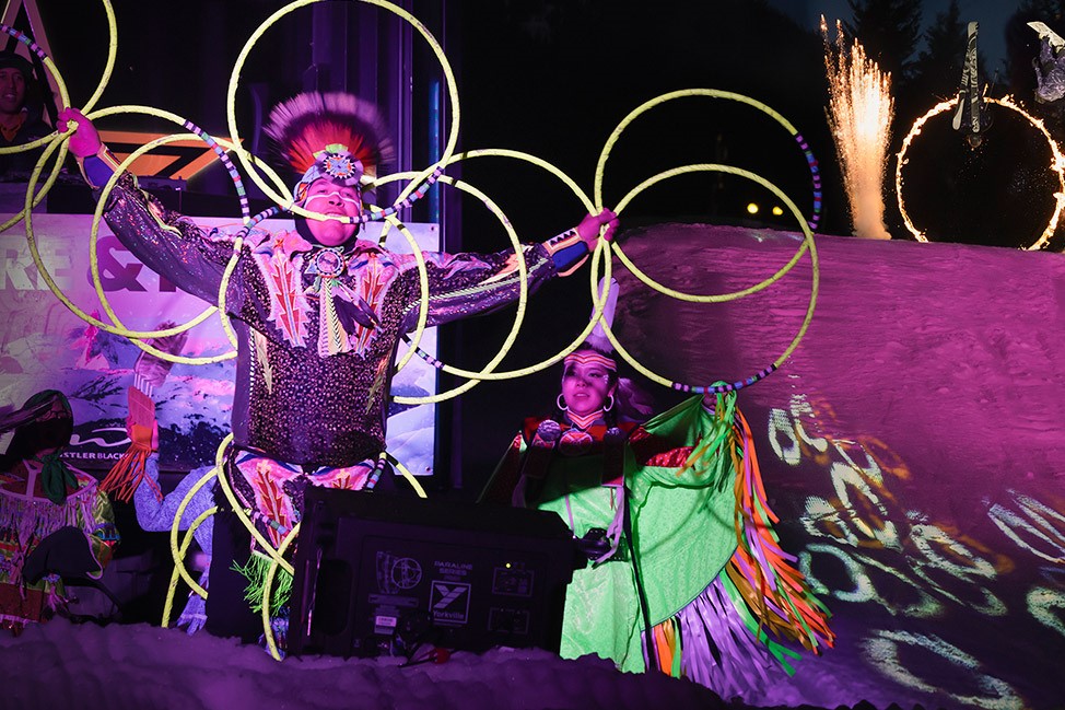 A performer dressed in colorful traditional attire showcases hoop dancing, surrounded by illuminated hoops, while another performer stands beside them on stage. The backdrop features vibrant lights and fireworks, creating a festive atmosphere.