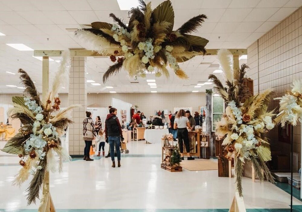 An event space featuring a floral arch made of dried plants and flowers, with people browsing stalls in the background. The setting is bright and spacious, indicating a gathering or market.