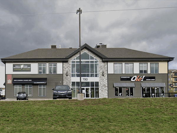 A modern two-story commercial building with large windows, flanked by a parking area and vehicles. The facade features a mix of stone and siding, set against a cloudy sky.