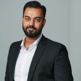 A professional-looking man with a beard wears a black suit and white shirt, standing confidently against a neutral background.
