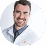 A smiling man in a lab coat and a collared shirt, standing against a plain background. He appears friendly, with a professional demeanor, suggesting a healthcare or medical setting.
