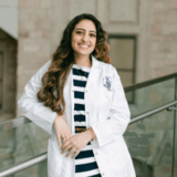 A smiling woman in a white lab coat stands confidently near a glass railing, dressed in a striped outfit. The background features light-colored stone walls, suggesting a professional or academic environment.