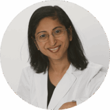 A smiling woman wearing glasses and a white lab coat, set against a plain background. She appears professional and friendly, suggesting a role in healthcare or medicine.