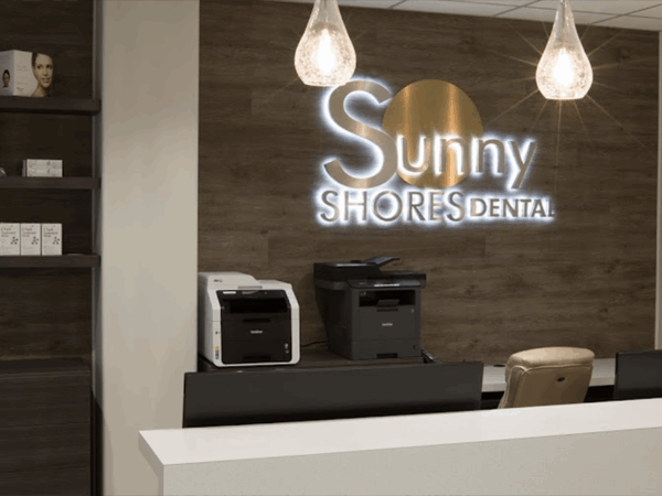 Reception area of a dental office with a front desk, a printer, and illuminated "Sunny Shores Dental" sign on a wooden wall. Three pendant lights hang from the ceiling.